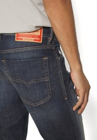 Mörkblå denimjeans med en röd etikett på baksidan, med kontrasterande orange sömnad och två bakfickor med en subtil V-formad design.