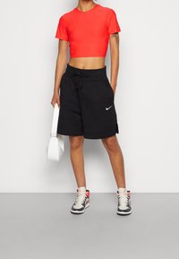 Röd kortärmad tröja med strukturerad mönster, svarta shorts med dragsko och en vit Nike-logotyp, matchat med högskor och en vit väska.