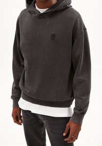 Tmavě šedá mikina s kapucí vyrobená z měkké tkaniny, která má vpředu klokaní kapsu a vyšívané logo na hrudi. Jednobarevná s volným střihem.