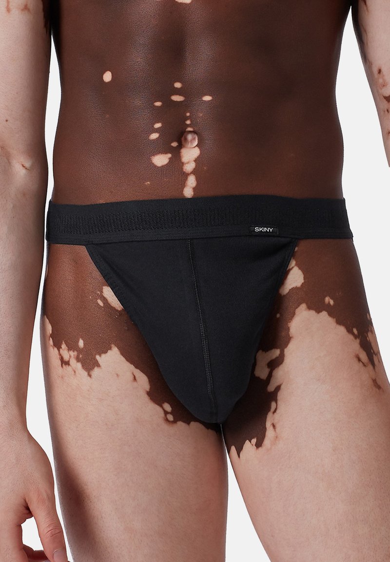 Svart tygstring med ett brett elastiskt midjeband, där varumärket "SKINY" syns fram. Modellens hud visar ljusa och mörka kontrasterande fläckar.