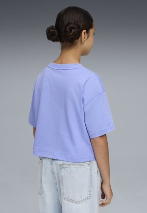 Persona joven con cabello oscuro recogido en dos moños, vistiendo una camiseta holgada lavanda y jeans azul claro, de pie y de espaldas sobre un fondo gris.