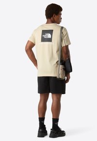 Muž v béžovém tričku The North Face, černých šortkách, černých ponožkách a botách, nese béžovou tašku přes rameno s lahví na vodu.