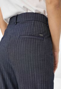 Pantalons rayés navy au coupe ajustée, avec une taille et une poche arrière. La texture est lisse avec de fines rayures verticales.