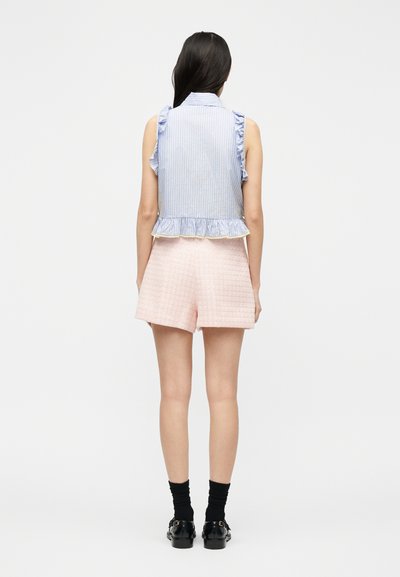 Γυναίκα που φαίνεται από πίσω φοράει αμάνικο μπλε ριγέ μπλουζάκι με βολάν, ροζ σόρτς ψηλόμεσο, μαύρες κάλτσες και μαύρα μοκασίνια σε λευκό φόντο.