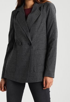 Kvinde iført en grå ternet dobbeltradet blazer med store lommer over en mørkerød top og sorte bukser.