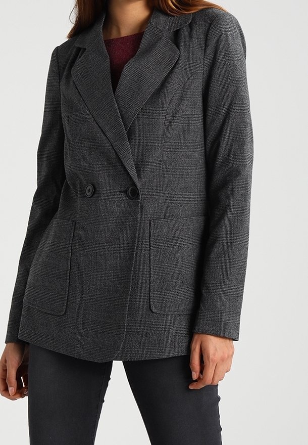 Femme portant un blazer à carreaux gris à double boutonnage avec de grandes poches, par-dessus un haut rouge foncé et un pantalon noir.