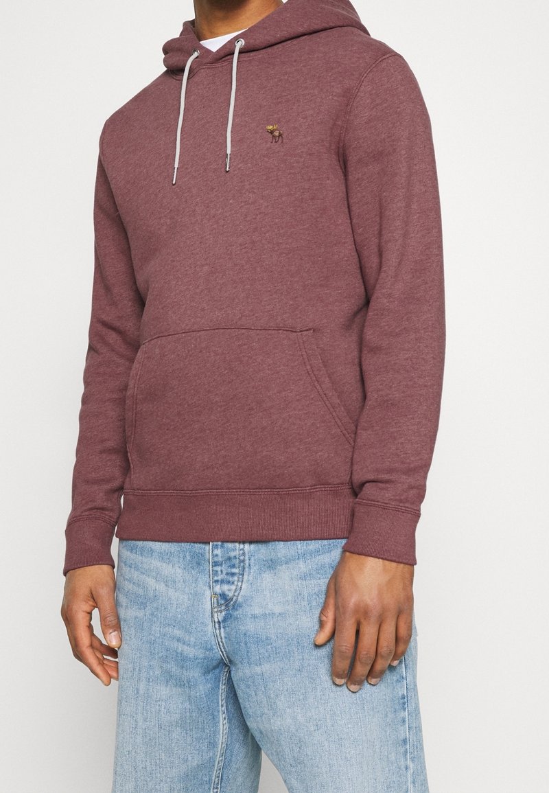 Uomo che indossa una felpa con cappuccio color bordeaux con coulisse bianche e piccolo logo moose ricamato, abbinata a jeans azzurri, in piedi su sfondo bianco.