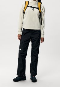 Valkoinen fleecepaita, mustat vedenpitävät housut ja keltainen reppu. Paidassa on korkea kaulus ja housuissa näkyvä logo. Mustat kengät.