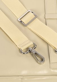 Dettaglio di una borsa sintetica beige con una tracolla testurizzata, un buckler in metallo argentato e un gancio, che mette in evidenza le cuciture e i dettagli in metallo lucido.
