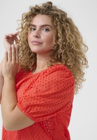 Haut rouge texturé avec un design perforé et des manches froncées. Le tissu semble léger, avec une silhouette ample.