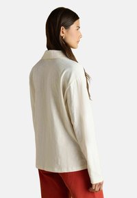 Naine, kellel on pikad pruunid juuksed, kannab lahtist kreemivärvi pikkade varrukatega pluusi ja punaseid pükse, seistes profiilis lihtsa tausta ees.