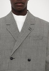 Dwurzędowa marynarka w czarno-białą kratę houndstooth, z dużymi klapami i dwoma czarnymi guzikami z przodu. Teksturowany materiał.