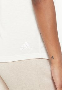 T-shirt bianco in cotone con colletto rotondo, dotato di un piccolo logo bianco Adidas vicino all'orlo. Lo sfondo presenta un tessuto beige.