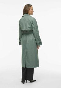 Trench coat lungo e verde, realizzato in materiale liscio, con cintura, revers ampi e spacchi laterali, abbinato a pantaloni neri e tacchi a punta.