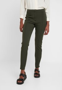 Olivově zelené společenské kalhoty s hladkou texturou, slim fit a bočními švy. V kombinaci s černými sandály na podpatku, oblečeno s lehkou blůzou.