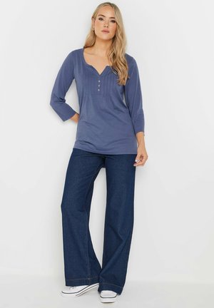 Haut à manches longues bleu foncé avec une patte de boutonnage et un design plissé, associé à un jean en denim foncé à jambes larges et des baskets blanches.