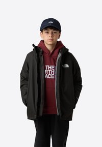 Černá nepromokavá bunda s kapucí, šedou podšívkou a logem The North Face. Nositelná přes vínovou mikinu s bílým logem, v kombinaci s tmavými kalhotami.