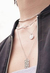 Trois colliers en argent à plusieurs couches : un pendentif rectangulaire texturé, un charme circulaire lisse et une chaîne simple. Porté sur un haut gris.