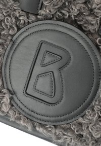 Étiquette en cuir gris avec la lettre "B" en relief sur un tissu bouclé gris texturé, comportant des détails de couture et une forme ronde.