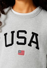 Šedá mikina se zvětšenými černými písmeny "USA". Obsahuje malou vyšívanou americkou vlajku pod textem. Materiál je měkká směs bavlny.