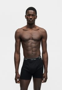 Άρτιος αρσενικός μοντέλο με σκούρο δέρμα που φοράει μαύρα μποξεράκια Calvin Klein, στέκεται μπροστά σε μια απλή ανοιχτό γκρι φόντο.