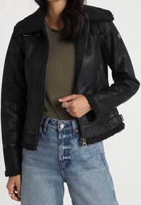 Czarna skórzana kurtka o teksturowanym wykończeniu, z kołnierzem i mankietami z owczej wełny, zapinana na zamek, w połączeniu z kontrastującymi jasnoniebieskimi dżinsami.