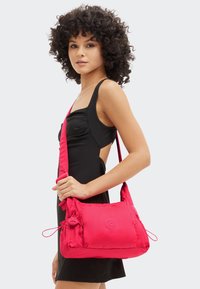 Rød nylon skuldertaske med justerbar rem, med en frontlomme, snorelukning og prægede logo. Glat tekstur, kompakt design.
