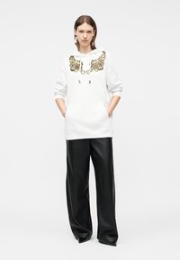 Femeie purtând un hanorac alb cu un model de tigru pe piept și pantaloni largi negri din piele, stând cu mâinile în buzunare pe un fundal alb.