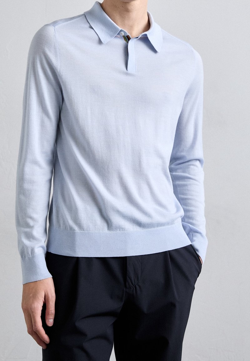 Homme portant un polo bleu clair à manches longues, rentré dans un pantalon noir, debout devant un fond uni gris clair.