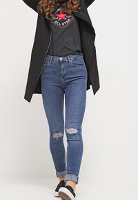 Jeans ajustados de mezclilla azul medio con rasgaduras desgastadas en las rodillas, combinados con un abrigo negro y una camiseta gráfica, completados con zapatillas negras.