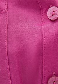 Camicia fucsia con bottoni, caratterizzata da una finitura liscia e texture; presenta un bottone rotondo e dettagli di cucitura lungo le cuciture.
