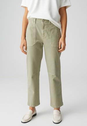 Personne portant un pantalon droit taille haute vert clair avec des poches plaquées à l'avant et des mocassins blancs à enfiler, sur un fond uni.