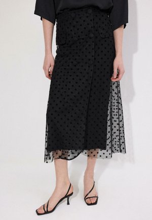 Μαύρη μίντι φούστα με πουά στρώση, λεπτομέρειες με κουμπιά και μπροστινές τσέπες, φορεμένη με μαύρα σανδάλια με λουράκια και τακούνι.