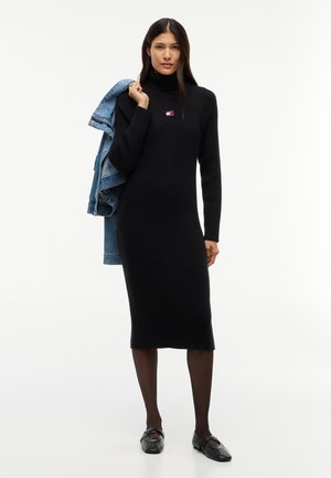 Vrouw draagt een zwarte geribde coltrui-jurk, zwarte panty's, zwarte schoenen, houdt een blauw spijkerjack over één schouder, staand op een witte achtergrond.