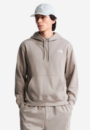 Jonge man draagt een beige North Face hoodie, bijpassende joggingbroek en een witte pet, handen in de zakken, kijkt opzij.