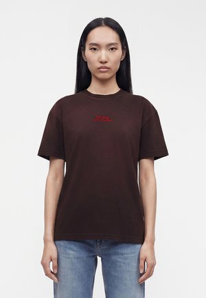 Kobieta o prostych czarnych włosach, ubrana w ciemnobrązowy T-shirt z czerwonym napisem i niebieskie dżinsy, stojąca na jednolitym białym tle.