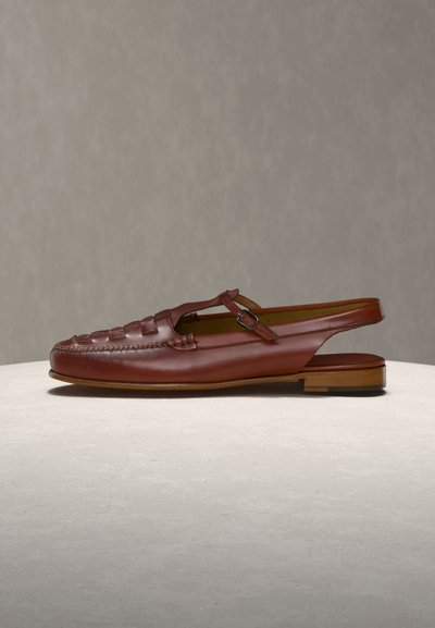 Καφέ δερμάτινο σανδάλι με πλεκτό σχέδιο στα δάχτυλα, λουράκι αστραγάλου με αγκράφα και χαμηλό ξύλινο τακούνι, τοποθετημένο σε ανοιχτή επιφάνεια με απλό φόντο.