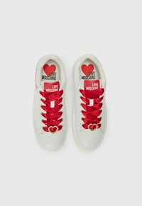 Weiße Sneakers mit roten Satinschnürsenkeln, goldfarbener, herzförmiger Hardware-Akzent und bedruckten Einlegesohlen, die "LOVE MOSCHINO" und ein Herzgrafik zeigen.