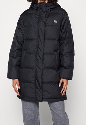 Manteau noir matelassé à capuche avec poches avant, porté sur un pantalon gris, mains partiellement visibles sur les côtés, fond blanc.