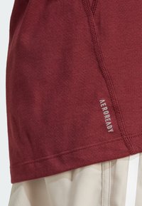 Camiseta deportiva color granate con tejido texturizado, con una costura lateral y la marca "AEROREADY" en gris. Corte entallado, mostrada con pantalones cortos de color claro.