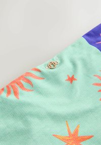 Közelkép zöld szövetről, amelyen narancssárga tengeri csillag és korall minták láthatók, valamint egy kis kerek arany fém címke varrva a szélénél.