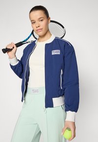 Blue bomber jacket with white accents and zipper pockets, paired with light green pants. Tennis racket and ball in hands.