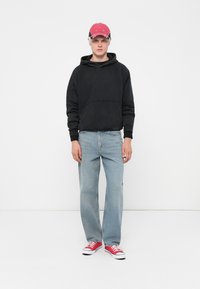 Svart hettegenser med kengurulomme, lyseblå rette jeans og røde sneakers. Capen er rød med logo-detalj. Enkel design, uformell passform.