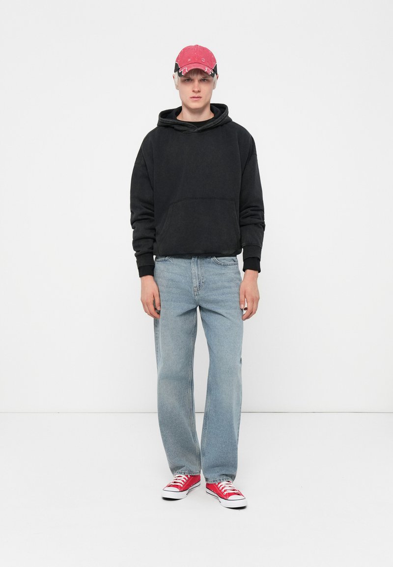 Felpa nera con cappuccio e tasca a marsupio, jeans dritti blu chiaro e sneakers rosse. Il cappellino è rosso con un dettaglio del logo. Design semplice, vestibilità casual.