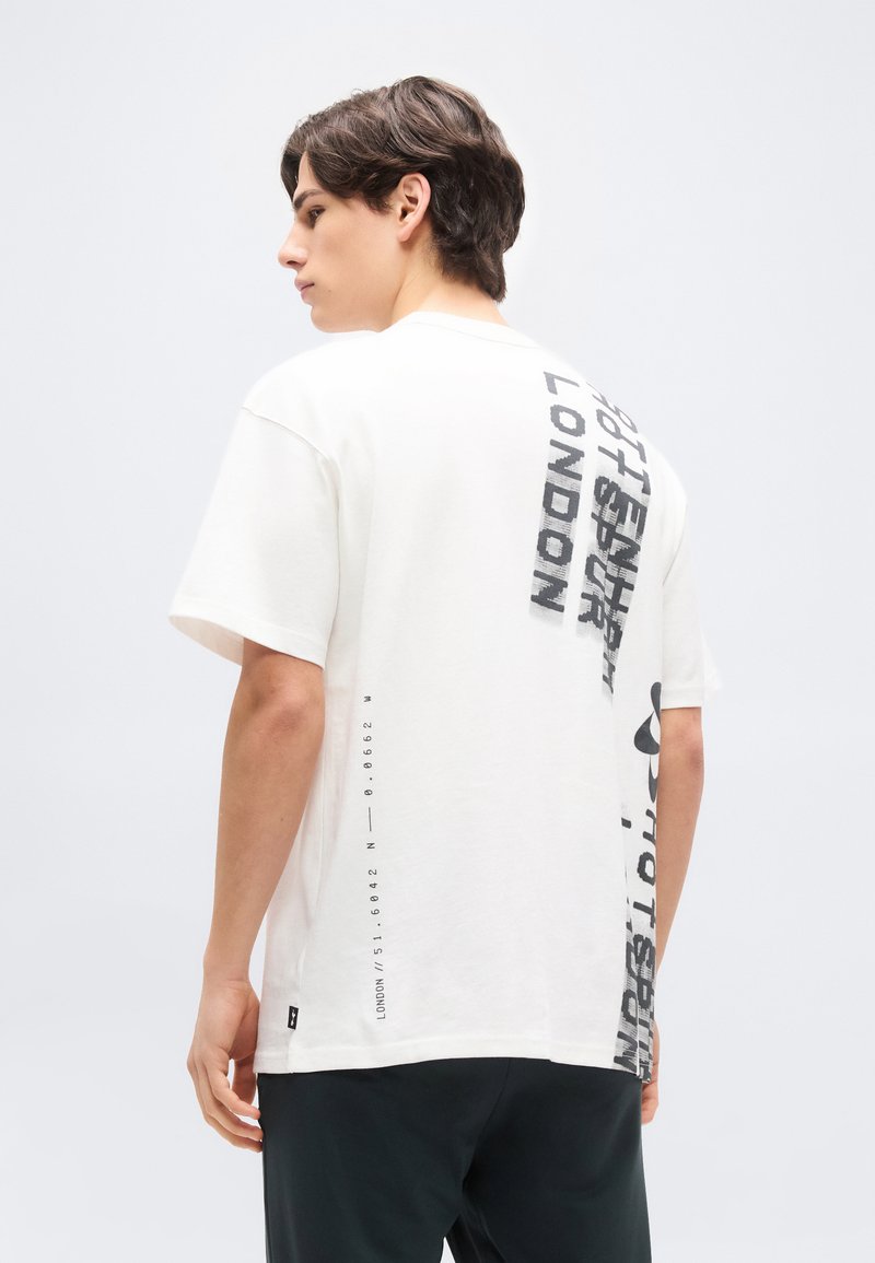 Νεαρός άνδρας με λευκό άνετο T-shirt με μαύρο κάθετο κείμενο στην πλάτη και μαύρο παντελόνι, στέκεται μπροστά σε απλό φωτεινό φόντο.