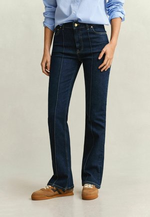 Personne portant un jean droit bleu foncé, une chemise bleu clair à manches longues retroussées et des baskets marron, debout sur un sol clair.