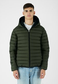 Giacca puffa verde oliva con design a trapuntatura orizzontale, zip frontale e cappuccio. Texture liscia e aspetto leggero, abbinata a jeans blu.