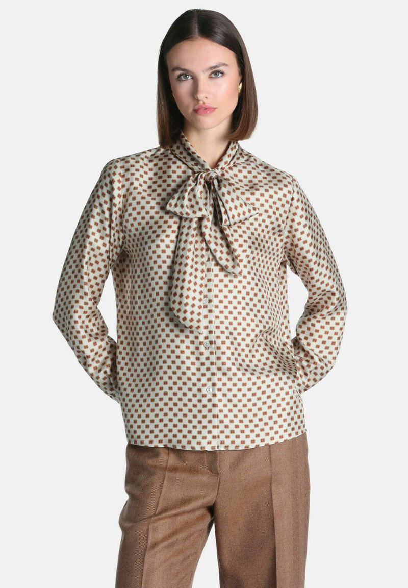Femme aux cheveux raides et foncés porte un chemisier à motifs avec un col à nœud papillon et un pantalon marron, debout les mains dans le dos sur fond blanc.