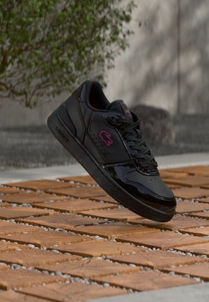 Black Lacoste sneaker with pink crocodile logo floating above wooden tile floor outdoors, blurred green plant and gray wall background.