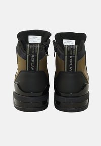 Zapatillas altas en verde olivo con acentos en negro, que cuentan con cierres de cremallera y forro textil, suela negra brillante y talón reforzado para mayor soporte.
